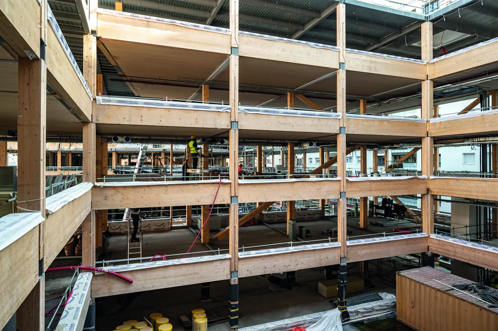 Int&eacute;rieur du b&acirc;timent en chantier : planchers en CLT et poteaux-poutres en lamell&eacute;-coll&eacute; sur plusieurs niveaux, avec des ouvriers