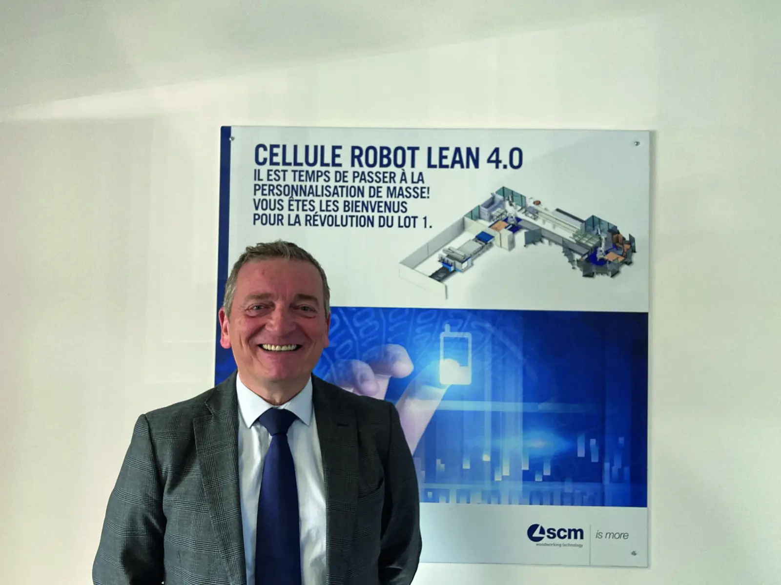 Laurent Mazi&egrave;s en costume devant une affiche SCM &laquo; Cellule robot Lean 4.0 &raquo;