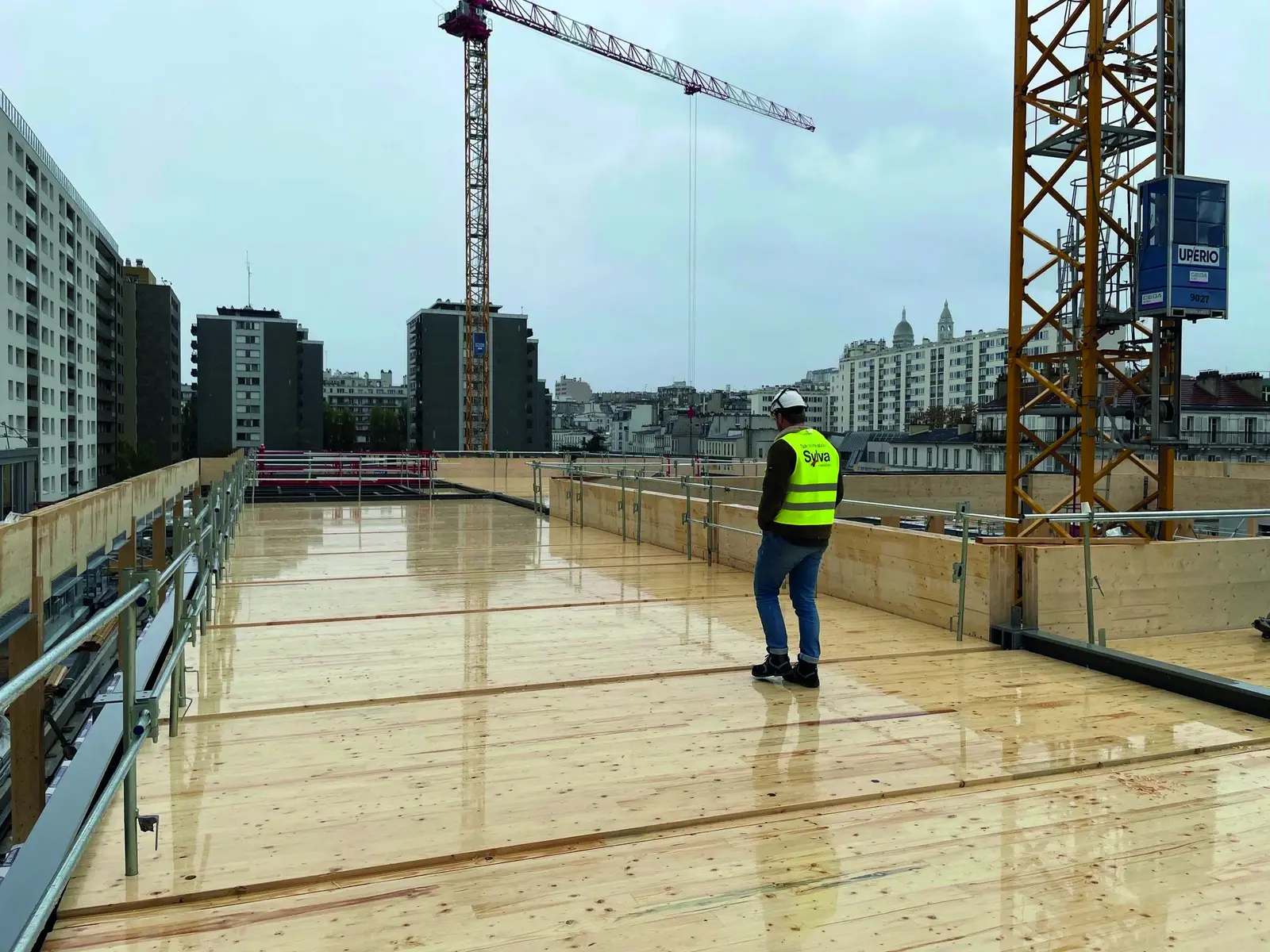 Ouvrier en gilet jaune sur un plancher en CLT au sommet du b&acirc;timent, grues en arri&egrave;re-plan